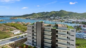 Un hôtel moderne à plusieurs étages, doté d’un jardin sur le toit et offrant une vue sur une ville côtière, avec des bateaux et des montagnes en arrière-plan.