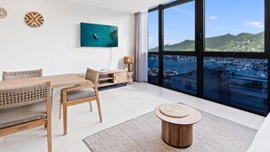 Un salon moderne avec un coin repas, de grandes fenêtres offrant une vue sur une ville côtière et un téléviseur fixé au mur qui diffuse une vue aérienne de la mer.