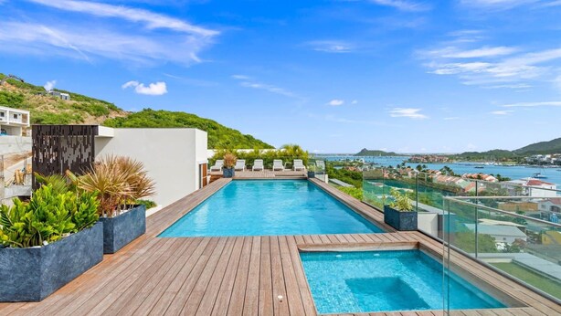 Ein Infinity-Pool auf dem Dach mit Holzterrasse, umgeben von Topfpflanzen und einem Glasgeländer mit Blick auf einen Yachthafen und die Skyline der Stadt.