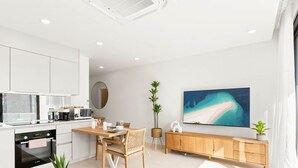 Une cuisine moderne avec des meubles blancs, un coin repas comprenant une table et des chaises en bois, un grand téléviseur à écran plat fixé au mur et un meuble en bois en dessous.