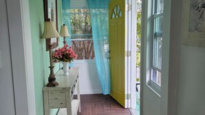 A hallway with a wooden floor, a white dresser with a lamp, a vase of flowers, a yellow door, and a window with a blue curtain. - Cozy Cute Beachy 2-bedroom house with Backyard, Parking & grill (Clearwater)