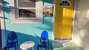 A small porch with two blue chairs and a white table, a yellow door, and a window with a view of a garden. - Cozy Cute Beachy 2-bedroom house with Backyard, Parking & grill (Clearwater)