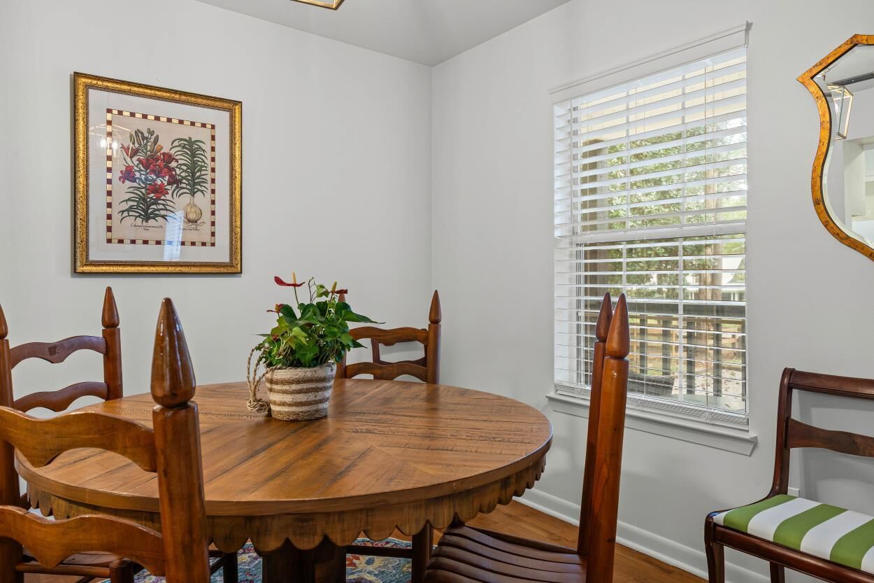 A dining room with a round wooden table, six wooden chairs, a potted plant, and a framed botanical illustration on the wall.