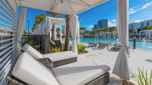 A covered outdoor seating area with white cushions and wicker furniture, overlooking a swimming pool with lounge chairs and umbrellas. - Beach Village Resort BVR07 | by Liquid Life (Orange Beach)