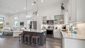 A modern kitchen with white cabinetry, a large island with stools, stainless steel appliances, and a view of the outdoors. - Beach Village Resort BVR07 | by Liquid Life (Orange Beach)