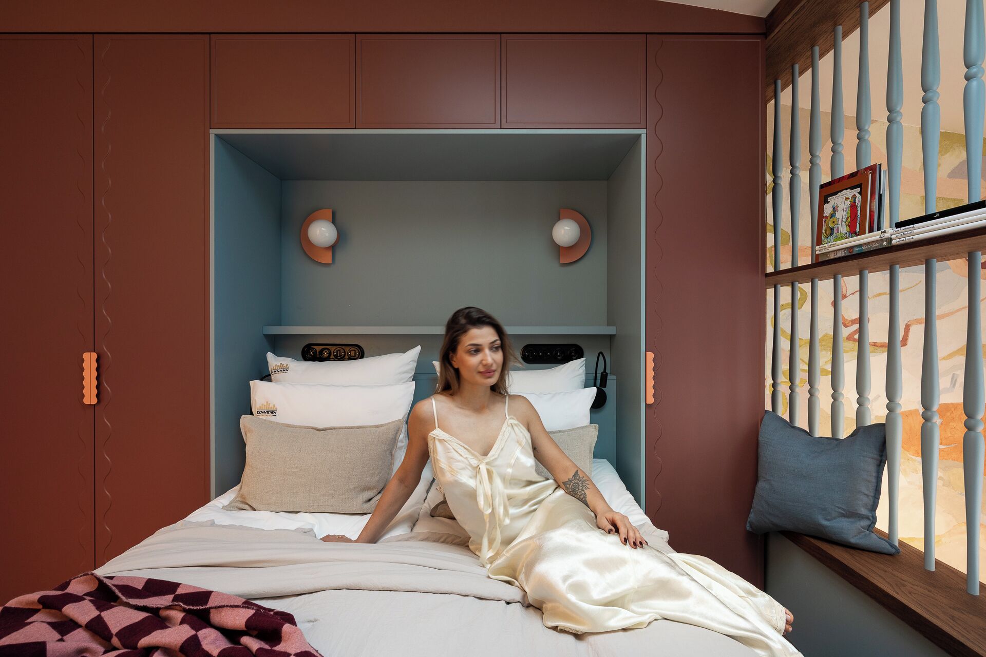 A bedroom with a bed, pillows, a nightstand, a shelf with books, and a window with blinds.