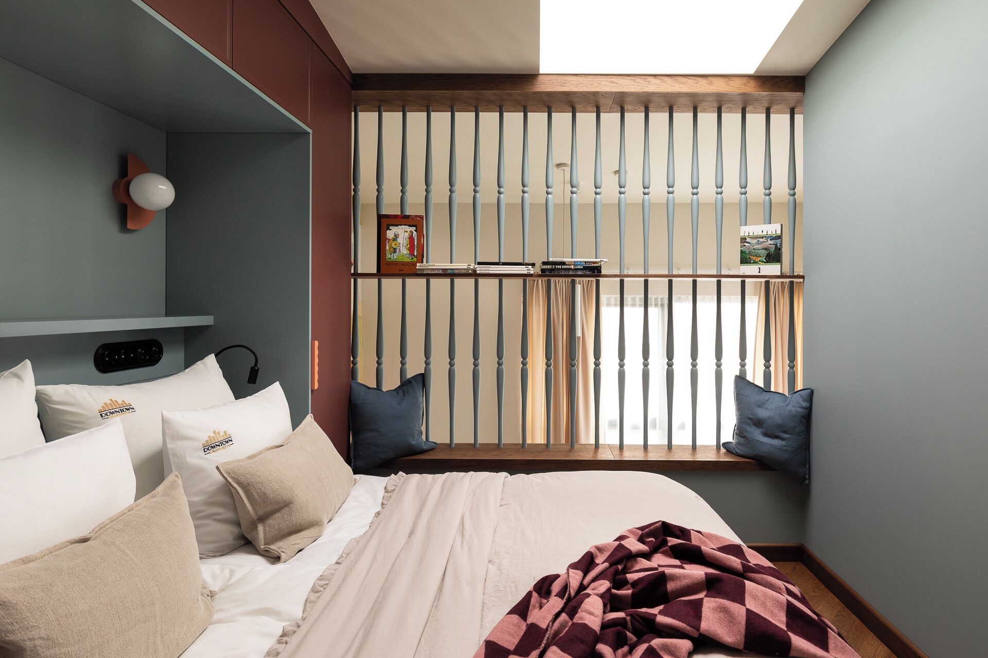 Een slaapkamer met een bed, kussens, een raam met jaloezieën, een plank met boeken en een wandlamp.