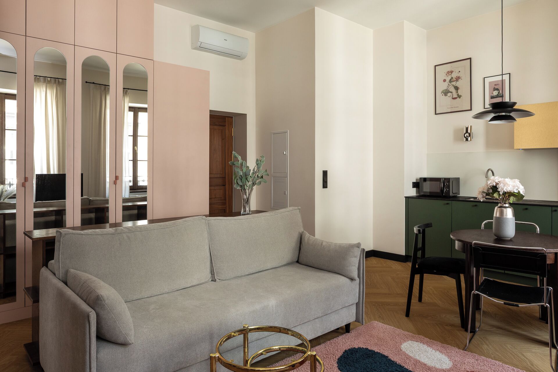 Ein modernes Wohnzimmer mit einem grauen Sofa, einem kleinen runden Tisch mit einer Vase sowie einer Küchenzeile mit Mikrowelle und Spüle.