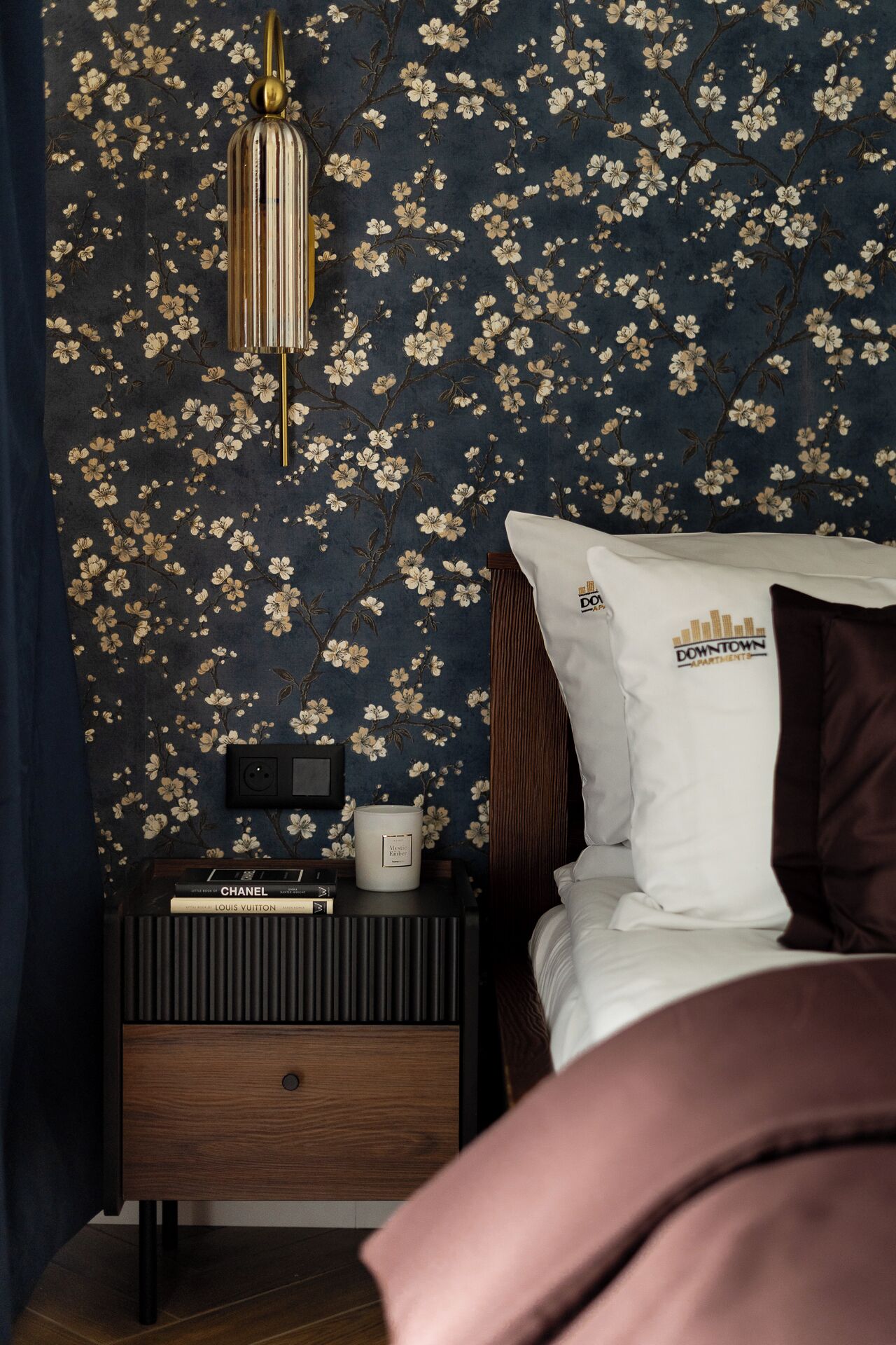 Une chambre à coucher avec un papier peint à motifs floraux sombres, une table de chevet avec des livres, une lampe et un lit avec un oreiller sur lequel est inscrit « Downtown Apartments ».