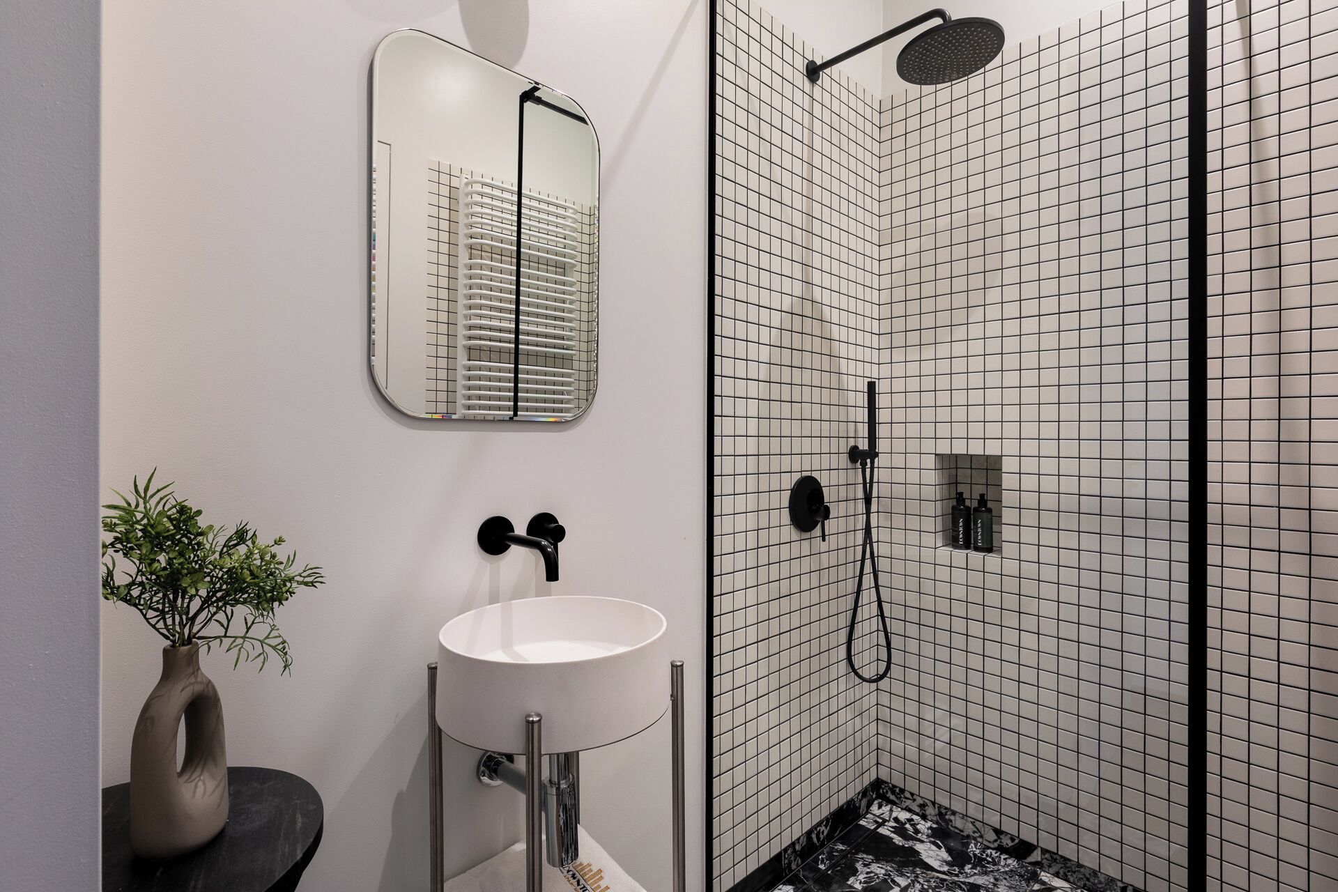 A modern bathroom with a white sink, a mirror, a shower with a black hose, and a tiled wall.