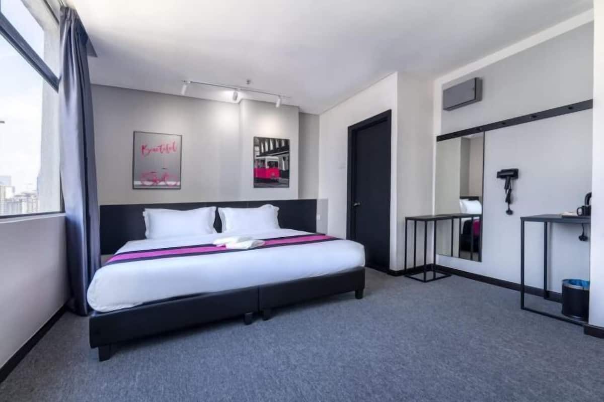 A hotel room with a large bed, a window with curtains, a mirror, and a small table with a chair.