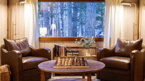 Ein gemütliches Zimmer mit zwei Ledersesseln, einem runden Holztisch mit Schachbrett, einem Bücherregal und einem Fenster mit Vorhängen.