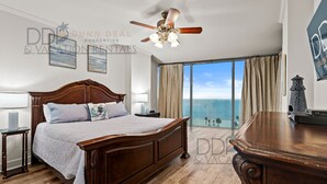 A bedroom with a large bed, a ceiling fan, a nightstand, a lamp, and a view of the ocean.