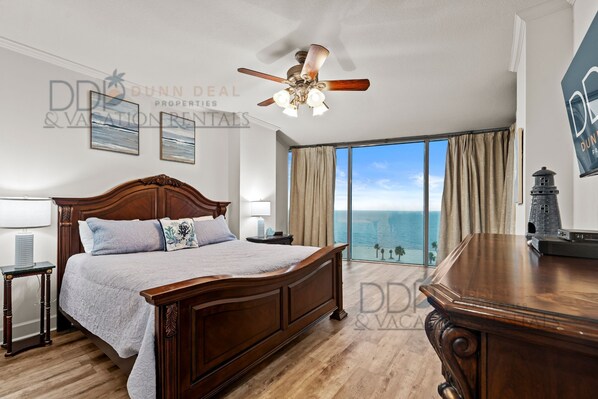 A bedroom with a large bed, a ceiling fan, a nightstand, a lamp, and a view of the ocean.