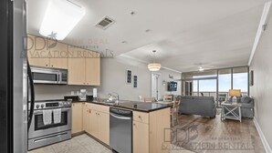 A modern kitchen with stainless steel appliances, a dishwasher, and wooden cabinets. There is a dining area with a gray sofa and a small table.