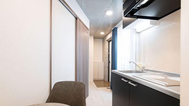 A narrow hallway with a kitchenette on the right, a chair in the foreground, and a door leading to another room. - Ferio Kamata II (Tokyo)