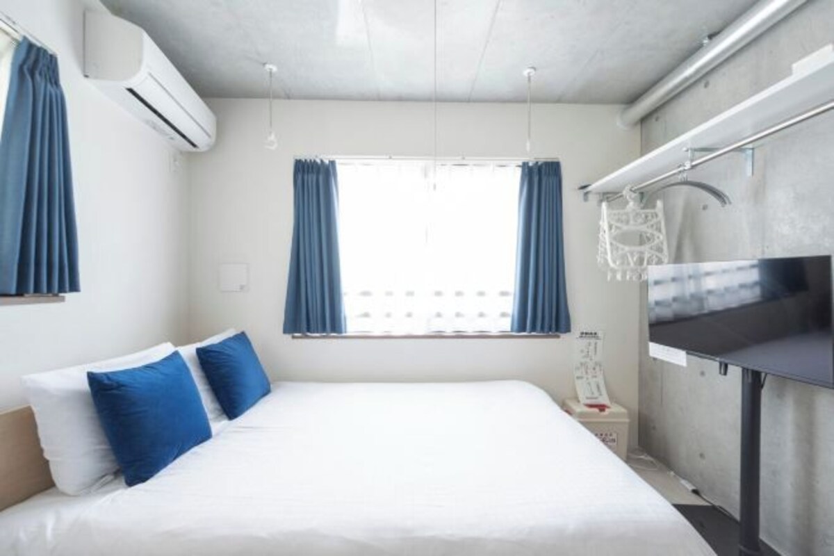 A modern hotel room with a large bed, a small bedside table, a window with curtains, and an air conditioning unit.