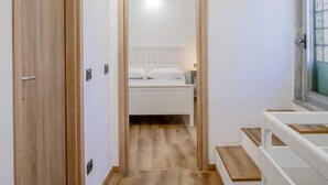 En smal hall leder in till ett sovrum med en säng, ett fönster och en trädörr.