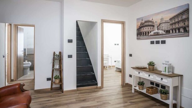 A modern interior with a staircase, a white console table with baskets, and a large painting of a historical building. - Profumo di Caruggi - Room Porto Antico (Genova)