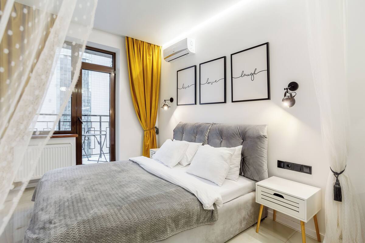 A bedroom with a bed, a bedside table, a wall-mounted air conditioner, and a window with a view of a balcony.