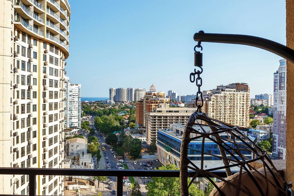 A balcony with a hanging chair, a view of a cityscape, and a building with balconies.