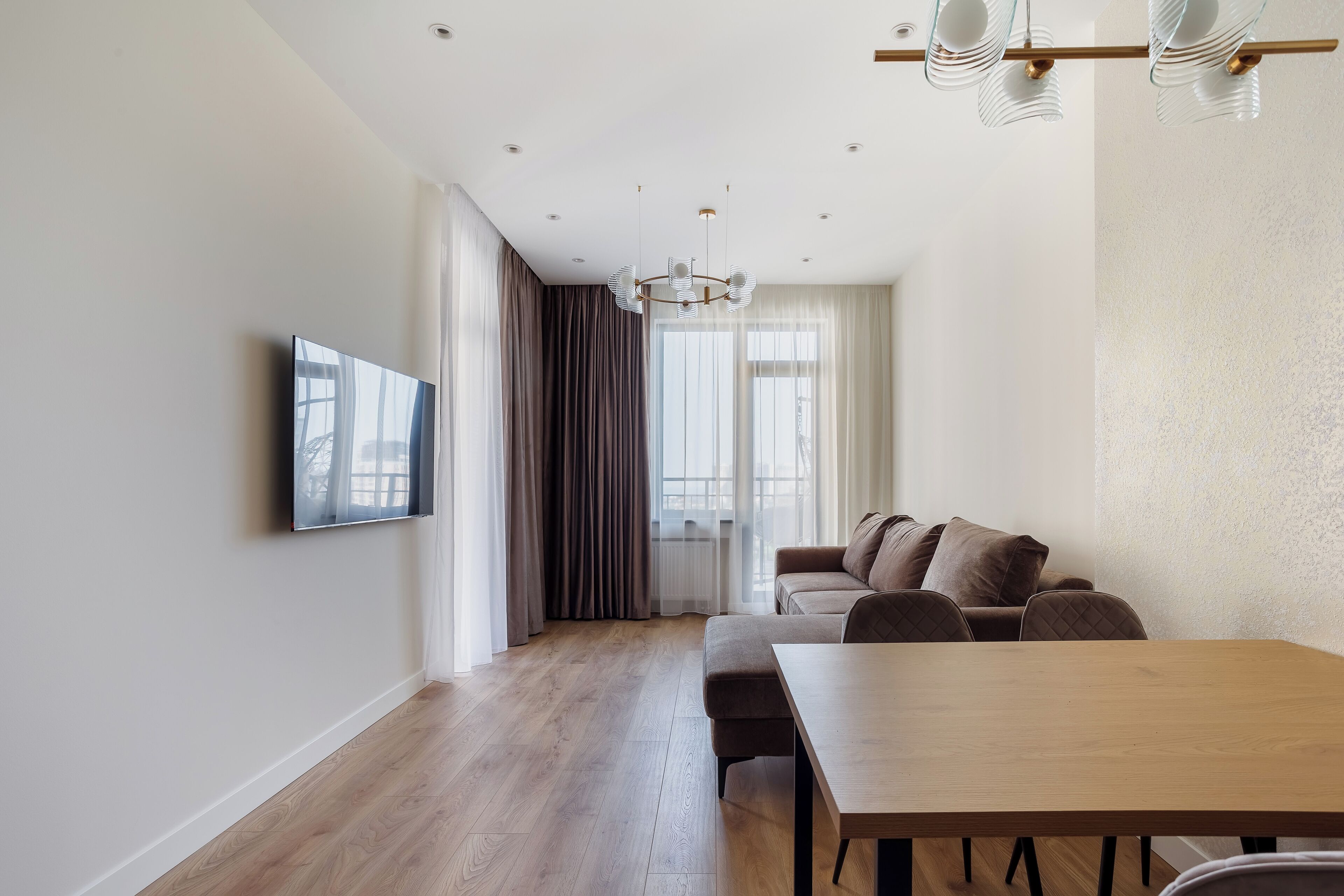 A modern living room with a wooden floor, a sofa, a dining table, and a TV mounted on the wall.