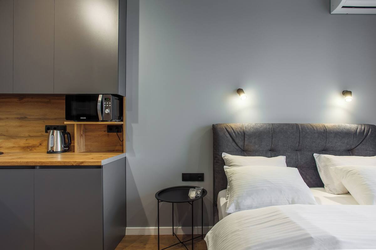 A modern hotel room with a kitchenette, a bed with white linens, and a grey upholstered headboard.