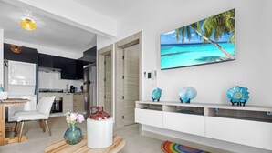 A modern kitchen with white cabinets, a dining area with a wooden table and chairs, and a living room with a white entertainment unit and a large beach-themed TV.