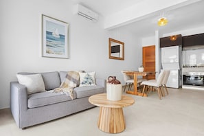Uma sala de estar moderna com um sofá cinzento, uma mesa de centro de madeira, uma área de jantar com uma mesa e cadeiras de madeira, uma cozinha com fogão e forno, e um quadro na parede.
