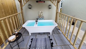 A small outdoor hot tub area with a wooden pergola, a patterned rug, and a small table with a bottle and a bowl. - Klysa (Camborne)