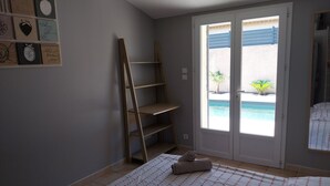 A bedroom with a bed, a wooden ladder, a window with a view of a pool, and a wall with framed pictures.