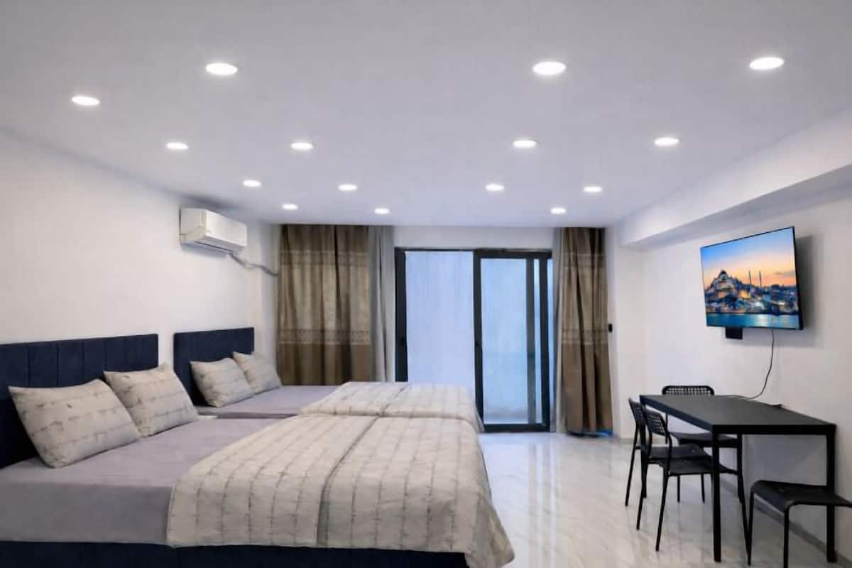 A hotel room with two beds, a dining table, chairs, a TV, and a large window with curtains.