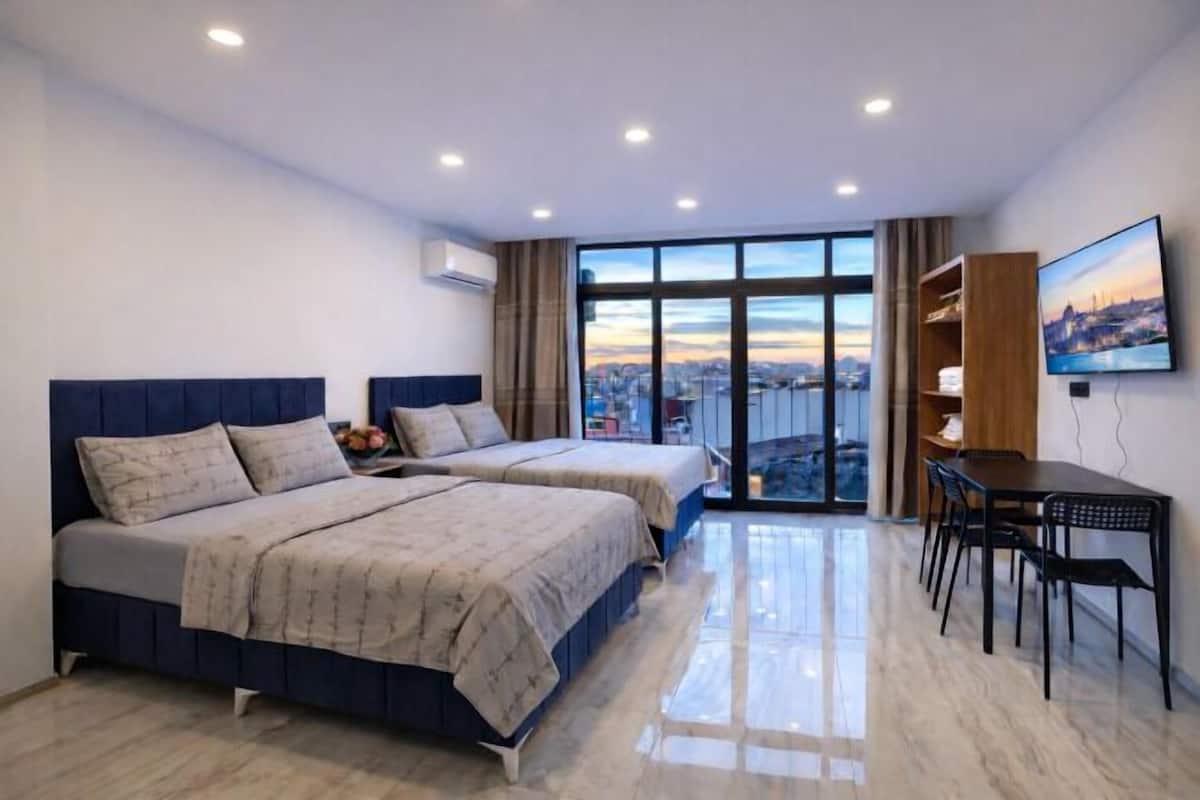 A modern hotel room with two beds, a dining area, and a large window offering a city view.