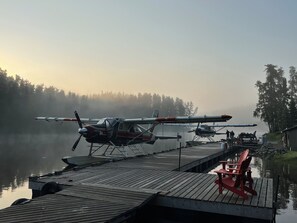 A seaplane docked at a wooden pier with a calm lake and forested hills in the background.