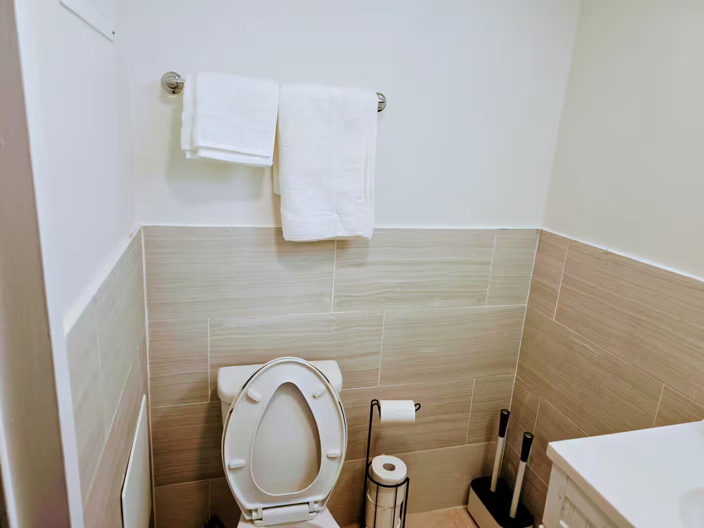 Un baño con inodoro, un perchero con dos toallas, dispensador de papel higiénico y un cepillo de limpieza.
