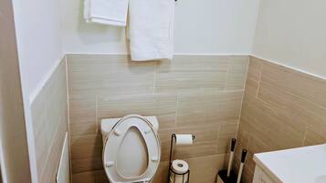 Un baño con inodoro, un perchero con dos toallas, dispensador de papel higiénico y un cepillo de limpieza.