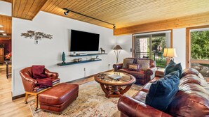 A spacious living room with leather furniture, a large TV mounted on the wall, and a view of the outdoors through large windows.