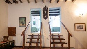 Una escalera con escalones y barandillas de madera, flanqueada por ventanas con contraventanas azules, un reloj de pared y cuadros enmarcados.