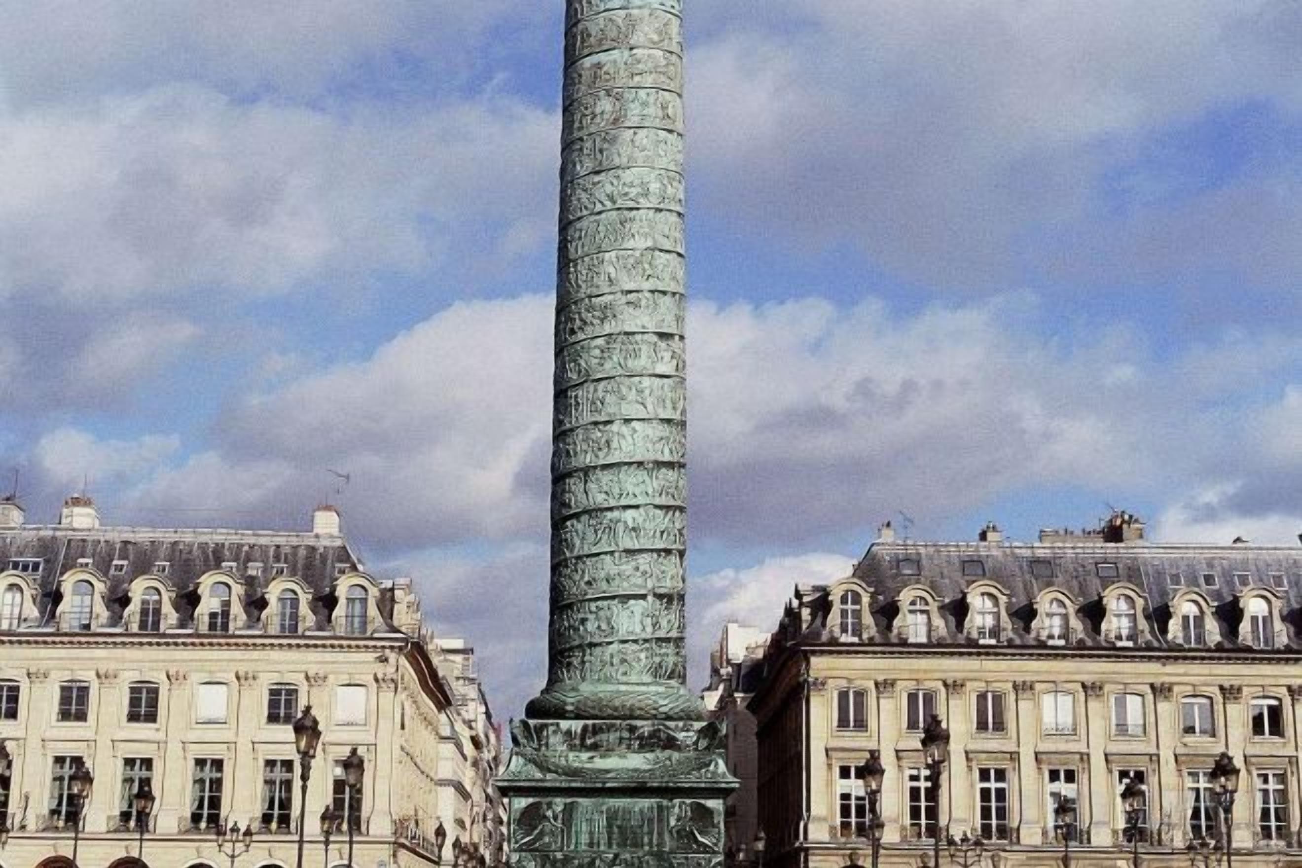 Une colonne haute et étroite, dont la texture est marquée par les intempéries, est entourée d’une architecture classique, avec de nombreuses fenêtres, et se dresse sous un ciel dégagé.