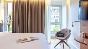 Una habitación de hotel con cama, una silla, un televisor y un ventanal con cortinas.