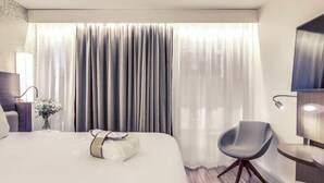 Ein Hotelzimmer mit einem Bett, einem Sessel, einem Schreibtisch, einer Lampe und einem großen Fenster mit Vorhängen.