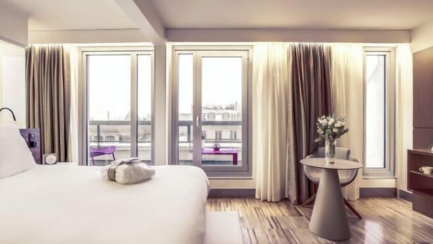 Una habitación de hotel con una cama grande, una mesa redonda y vistas a edificios a través de las ventanas.