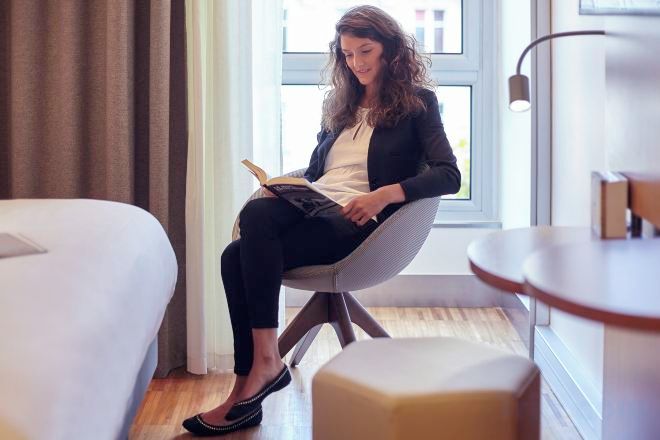 Una persona sentada en una silla leyendo un libro en una habitación con una cama, una mesita y una ventana.