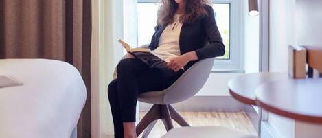 Una persona sentada en una silla leyendo un libro en una habitación con una cama, una mesita y una ventana.