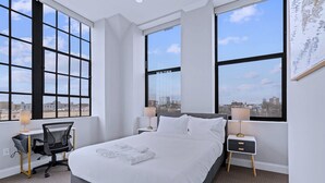 Een moderne hotelkamer met een groot bed, twee nachtkastjes, een bureau met stoel, en grote ramen met uitzicht over de stad.