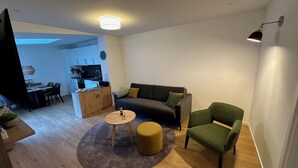 A modern living room with a dark grey sofa, a green armchair, and a round wooden coffee table. A kitchen with white cabinets and a dining area with a table and chairs is visible in the background. - Apartment Bergblick | Top vacation apartment with view of mountains and Ahr 1-4 persons (Altenahr)