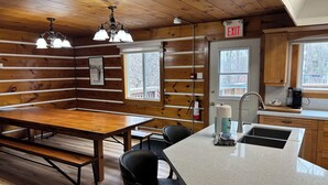 Een chaletinterieur met een houten eethoek, zwarte stoelen, een keuken met spoelbak en koffiezetapparaat, en een vluchtwijzer.