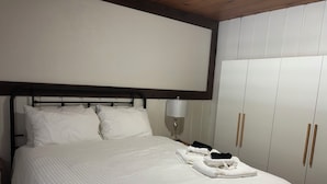 Een net opgemaakt bed met witte lakens en kussens, een bedlampje en een garderobe met houten handgrepen.