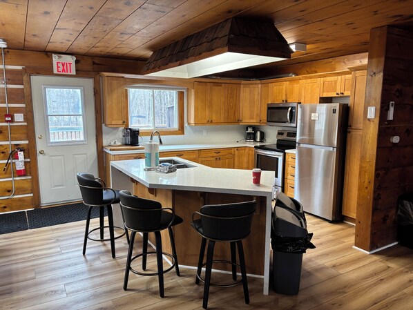 A cabin-style kitchen with wooden cabinets, a central island with bar stools, a microwave, a refrigerator, and an exit sign. - Blue Mountain Chalet ( 7 Bedrooms, Sleeps 15 Fully air-conditioned throughout . (town of Blue mountains)