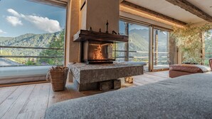 A stone fireplace with a fire, large windows, and a view of mountains.